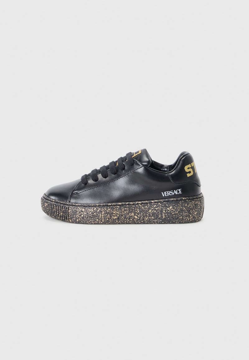 Zapatilla de cuero negro con suela texturizada salpicada de oro. Presenta cordones negros, un logotipo sutil en el lateral y un diseño de puntera redondeada.