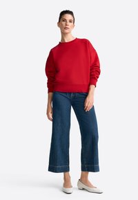 Červená mikina s okrúhlym výstrihom a dlhými rukávmi, skombinovaná s širokými tmavomodrými džínsami a bielymi balerínkami. Hladký materiál a uvoľnený strih.