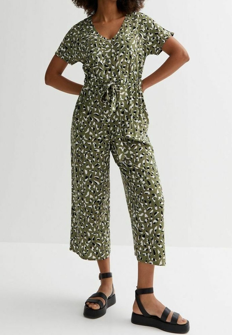 Femme portant une combinaison kaki à motifs à manches courtes et des sandales compensées noires, debout, les mains sur les hanches.
