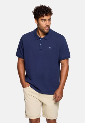 Hombre con cabello rizado, vestido con una camiseta polo azul marino y pantalones cortos beige, de pie con una mano en el bolsillo, mirando hacia un lado.