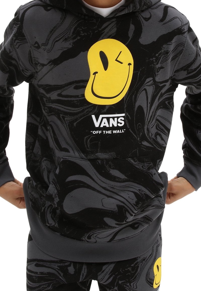 Sweat à capuche noir marbré avec un smiley jaune qui fait un clin d'œil et le logo blanc Vans "Off the Wall" sur la poitrine, porté par une personne.