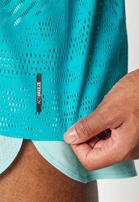 T-shirt de sport turquoise avec texture en mailles, présentant des perforations circulaires. Étiquette de logo "ACTIBREEZE" près de l'ourlet. Superposé avec un short léger bleu.