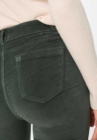 Mørkegrønne jeans med en glat tekstur. Har en baglomme med en spids form, diskrete syninger og en metalbrandetiket.