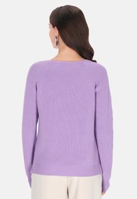 Lila kötött pulóver raglán ujjakkal, bordázott textúrával és lekerekített nyakkivágással. Egyenes alsó szegéllyel rendelkezik, és világos színű nadrág fölött viselik.