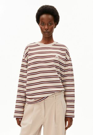 Giovane donna con capelli corti e ricci che indossa una camicia crema a maniche lunghe con righe orizzontali marroni e pantaloni beige chiaro.