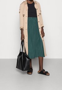 Trench coat beige con bottoni, top nero, gonna plissettata verde, borsa a tracolla nera e sandali scuri. Focus sui strati e sul contrasto dei colori.