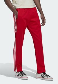 Red athletic pants featuring white side stripes, elastic waistband, and tapered fit, made from a smooth, lightweight fabric.