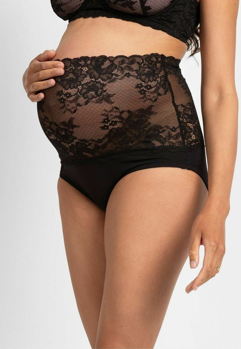 Tehotná žena v čiernom tehotenskom prádle z čipky s vysokým pásom, jemne drží bruško jednou rukou.