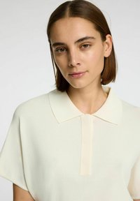 Polo à manches courtes en blanc cassé, fabriqué dans un tissu doux et léger, avec un col classique et une patte de boutonnage minimaliste.