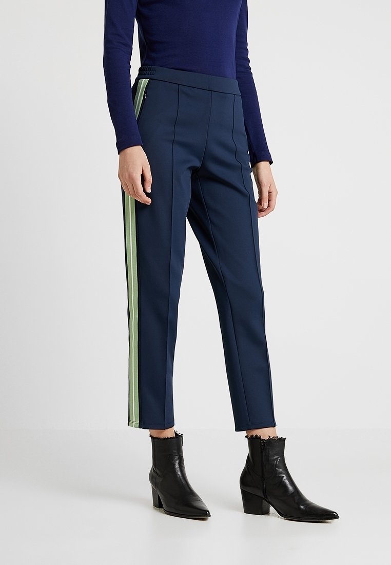 Pantalon bleu marine sur mesure avec des bandes latérales vertes. Fabriqué en tissu lisse, il présente une taille haute et une coupe slim raccourcie. Porté avec des bottines noires.