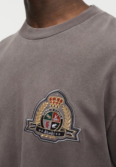 Γκρι βαμβακερό t-shirt με στρογγυλή λαιμόκοψη και περίτεχνο κεντημένο λογότυπο με χρυσές λεπτομέρειες και κορώνα, τοποθετημένο στην αριστερή πλευρά του στήθους.