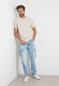 T-shirt beige in cotone a maniche corte, abbinato a jeans cargo blu chiaro con tasche laterali, indossato con sneaker bianche.