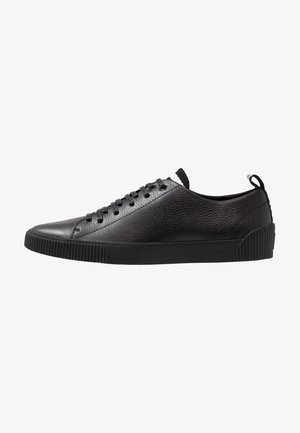 Schwarzer Sneaker mit niedrigem Profil aus Leder, strukturiertem Obermaterial, schwarzer Gummisohle, schwarzen Schnürsenkeln und Zuglasche an der Ferse, seitliche Ansicht.