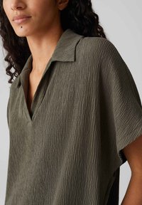 Short-sleeve, olive green shirt with a textured fabric, featuring a collar and a v-neck design, visible fine lines pattern throughout.