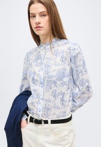 Νεαρή γυναίκα ντυμένη με μπλε και λευκό πουκάμισο με σχέδια, κρατώντας ένα σκούρο μπλε σακάκι, με το ένα χέρι στην τσέπη του κρεμ παντελονιού.