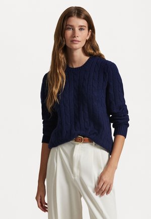 Navy cable-knit sweater with a round neckline and ribbed cuffs, paired with light-colored trousers and a brown belt. Soft texture and relaxed fit.