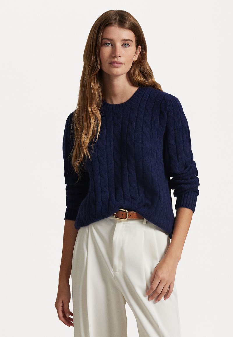 Pull en maille torsadée navy avec un col rond et des poignets côtelés, associé à un pantalon de couleur claire et une ceinture marron. Texture douce et coupe décontractée.