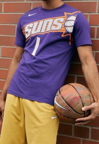 Lilla bomulds-t-shirt med "Suns" grafik og nummer "1," kombineret med gule shorts, holder en farverig basketball. Nike-logoer synlige.