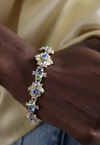 Gullfarget armbånd med blå og grønne edelstener i blomsterformer, med klare detaljer og et detaljert design, båret på et mørkhudet håndled.