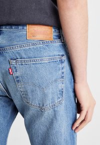 Jeans de mezclilla azul con un parche de cuero marrón etiquetado "501" en la parte trasera de la cintura, con dos bolsillos traseros y detalles de costura en naranja.