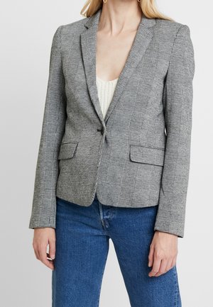 Femme portant un blazer gris à carreaux ajusté par-dessus un haut en maille blanc et un jean bleu, debout devant un fond clair uni.