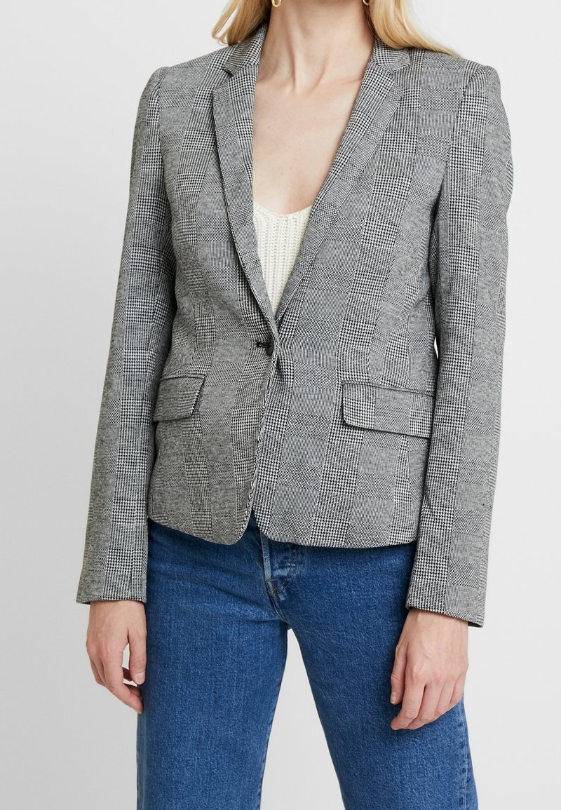 Femme portant un blazer gris à carreaux ajusté par-dessus un haut en maille blanc et un jean bleu, debout devant un fond clair uni.