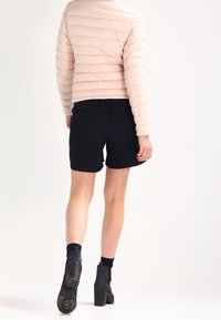 Gepolsterte, hellrosa Jacke mit hohem Kragen, kombiniert mit schwarzen Shorts und Stiefeletten. Glatte Textur und horizontales Steppmuster.