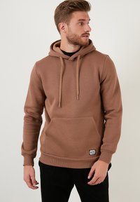 Sudadera marrón de tela suave, con un bolsillo frontal, cordones ajustables y puños acanalados. Etiqueta decorativa en el dobladillo.