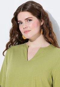 Olijfgroen top met een V-hals ontwerp, korte mouwen en een gladde textuur. Accessoires zijn gele, geweven hoop oorbellen.
