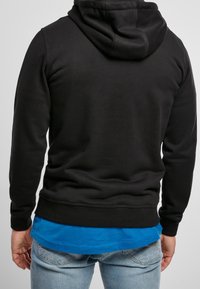 Czarna bluza z kapturem, wykonana z miękkiego, teksturowanego materiału, z kieszenią typu kangur i kapturem. Nosi się ją na niebieskiej koszuli i w parze z jasnoniebieskimi dżinsami.