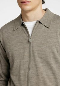 Siv zgoraj zapet pulover s ovratnikom, ki ima gladko teksturo in subtilne pletene detajle. Obut je čez belo srajco.