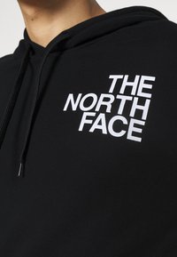 Černá mikina s kapucí s velkým bílým logem "THE NORTH FACE" na přední straně. Materiál vypadá texturovaně s nastavitelnými šňůrkami v kapuci.