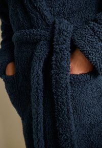 Mørkeblå badekåbe med blød, fluffy tekstur, bindebælte i taljen og to forlommer. Stoffet virker blødt og varmt, ideelt til afslapning.