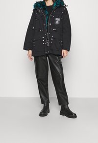 Svart parka-jacka med en teal faux fur-huvudbonad, flera framfickor, dragsko i midjan och silverfärgade detaljer. Bärs med svarta läderbyxor.