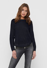 Pull bleu marine avec un motif texturé, manches longues et col rond, associé à un jean gris ajusté.