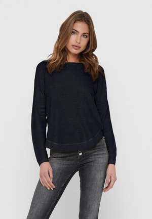 Pull bleu marine avec un motif texturé, manches longues et col rond, associé à un jean gris ajusté.