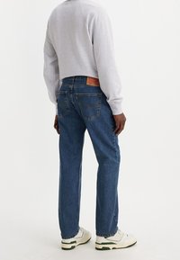 Man med blå raka jeans, ljusgrå långärmad tröja och vita sneakers som står mot en vit bakgrund.