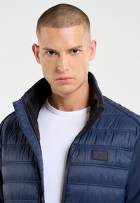 Jūras zilas krāsas pūšamā veste ar izšūtu rakstu, augstu apkakli un melniem akcentiem. Uz krūšu daļas ir zīmola birka un gluda iekšējā odere.