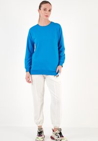 Sudadera azul brillante de manga larga y cuello redondo; combinada con pantalones deportivos blancos con puños que presentan un detalle bordado en negro. Zapatillas deportivas.