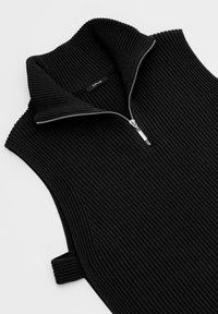 Gilet en maille noir côtelé sans manches avec un col montant replié et une fermeture éclair sur le devant, avec un petit détail de boucle latérale.