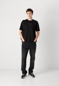 Black cotton t-shirt with logo on the front, paired with dark denim jeans, and black sneakers with white accents. Model standing with hands in pockets.