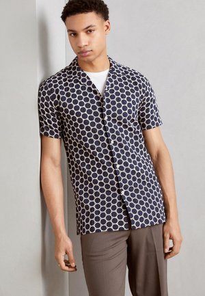 Chemise à manches courtes bleu marine avec un motif de cercles géométriques blancs, dotée d'un col et d'une fermeture à boutons, assortie à un pantalon marron.