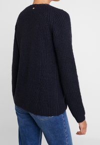 Maglione lavorato a maglia color blu notte scuro con finitura testurizzata, scollo rotondo e spalle scese. Presenta un piccolo accento dorato sul retro.