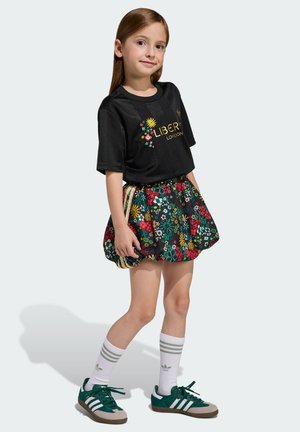 Tricou negru cu mâneci scurte, cu text auriu „LIBERTY” și design floral, asortat cu o fustă colorată cu model floral și adidași verzi.