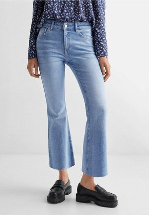 Kvinde iført lyseblå cropped flare-jeans, sorte chunky loafers og en blå blomstret bluse med lange ærmer, stående mod en ensfarvet baggrund.