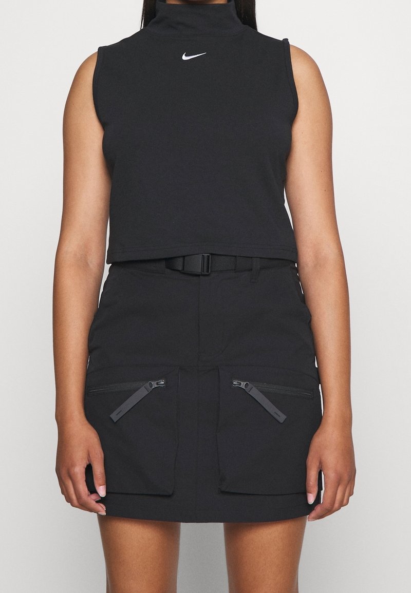 Svart kortärmad top utan ärmar med hög hals och vitt Nike-logotyp, i kombination med en svart kjol med två dragkedjefickor och ett bälte.
