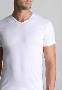 T-shirt bianca in cotone con scollo a V e maniche corte, caratterizzata da una texture liscia e un design aderente, mostrata su uno sfondo grigio chiaro.