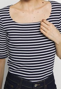Námořnické a bílé pruhované tričko se čtvercovým výstřihem a krátkým rukávem, vyrobené z měkké látky. Modelka má na sobě džíny v modré barvě se zlatým pro�šíváním.