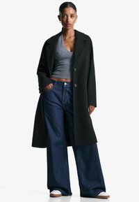 Zwarte wollen jas over een grijze cropped tanktop, gecombineerd met wijde blauwe denim jeans en witte sneakers. Heeft grote knopen en zijzakken.