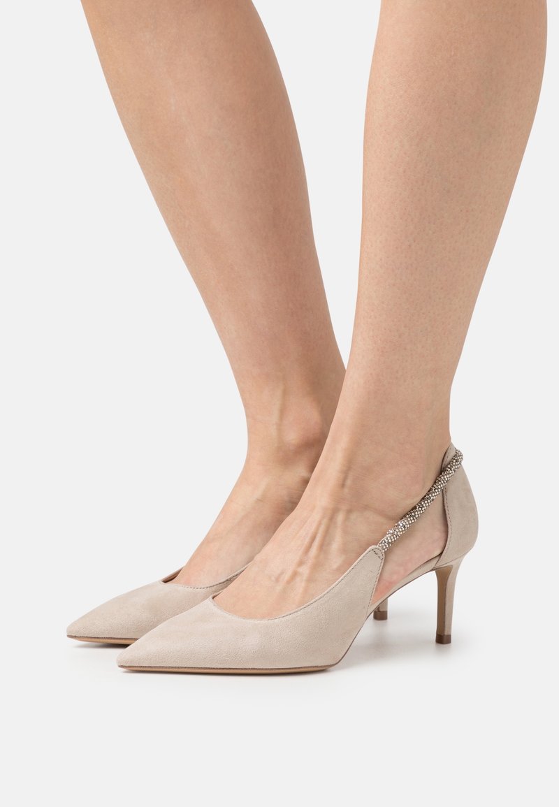 Femme portant des escarpins slingback en daim beige à bout pointu, avec des talons aiguilles fins de hauteur moyenne et une bride perlée autour de la cheville.